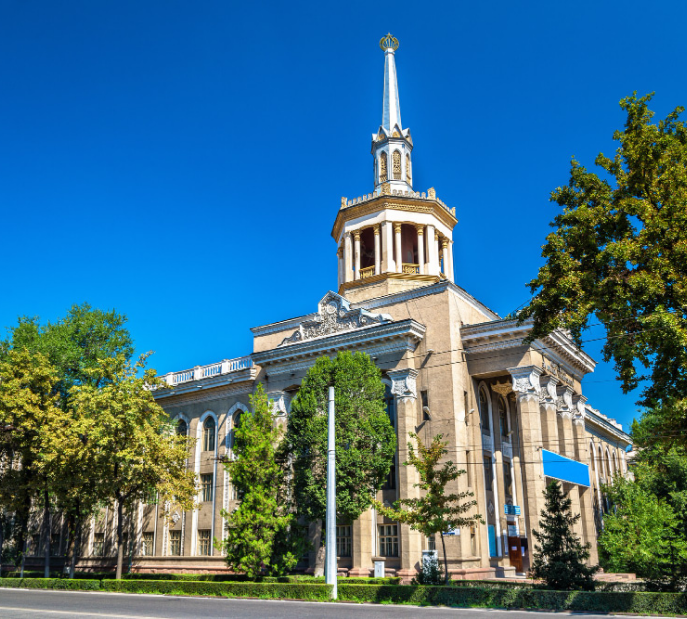 Large historic building with columns and a tall spire surrounded by trees on a sunny day.